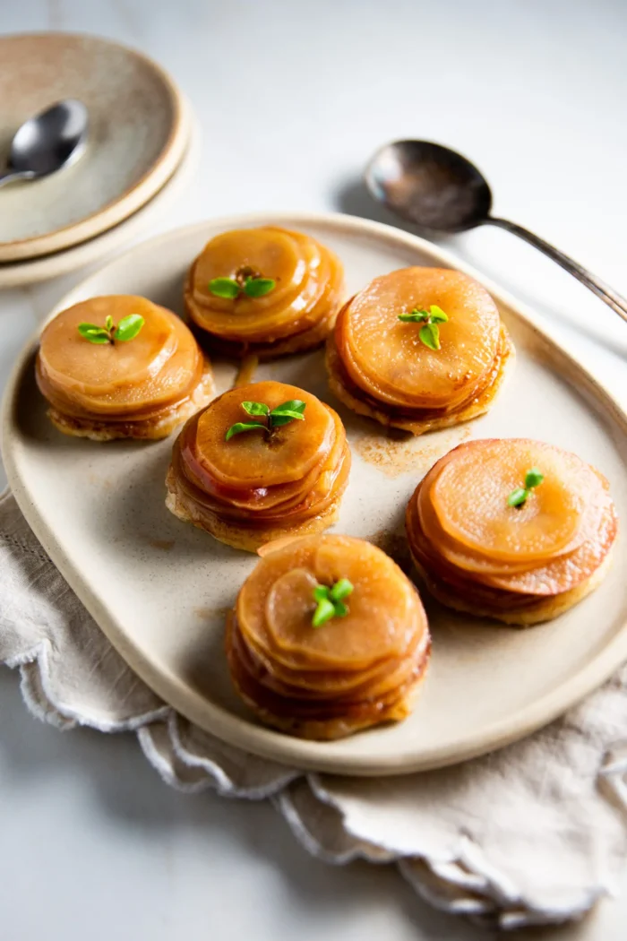 A beige plate holds six neatly stacked Mini Apple Tarts made of glossy, caramelized apple slices, each garnished with a green herb sprig. The plate rests on a folded cloth, with a large spoon and stacked dishes in the background.