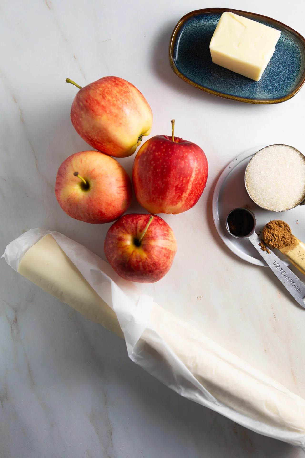 Four red apples, a blue plate with butter, sugar in a measuring cup, pie dough wrapped in white paper, and spoons with vanilla and cinnamon are arranged on marble—everything you need for this easy Mini Apple Tarts muffin-tin recipe.