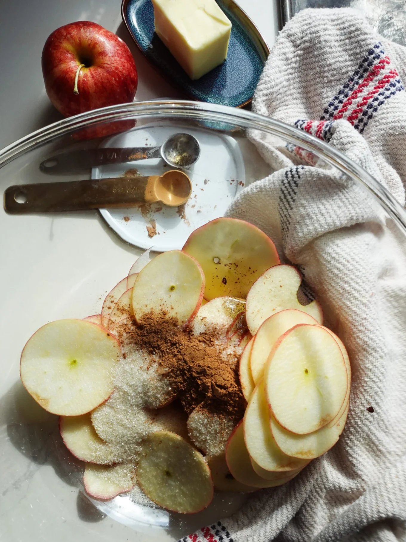 A glass bowl holds thinly sliced apples for Mini Apple Tarts, topped with cinnamon and sugar. Nearby are measuring spoons, butter on a dish, a red apple, and a striped towel on a sunlit white countertop—perfect for your next muffin-tin recipe.