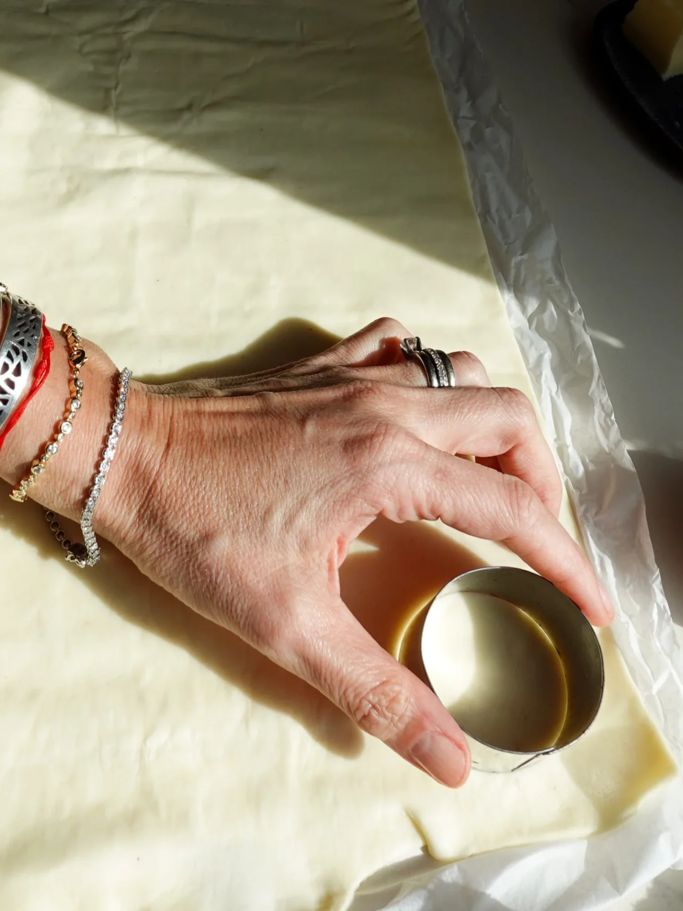 A hand with bracelets and rings presses a round metal cookie cutter into pastry dough on parchment paper in bright natural light—prepping for easy Mini Apple Tarts, the perfect muffin-tin recipe for any occasion.