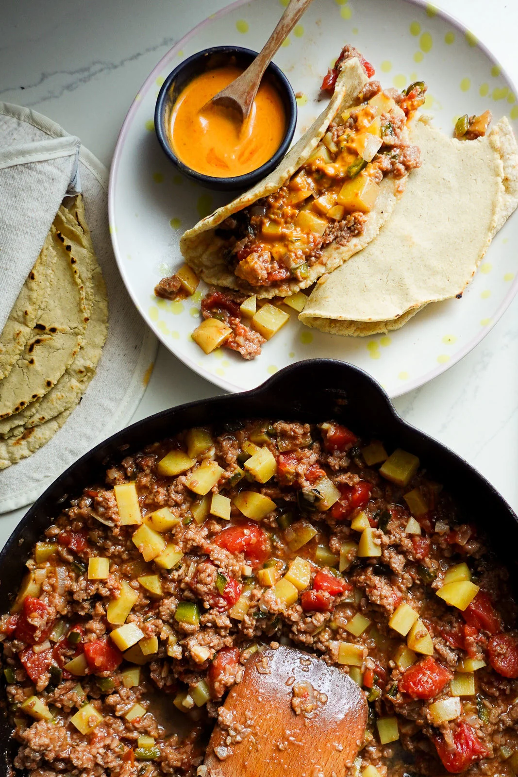 A skillet filled with ground beef, diced potatoes, tomatoes, and vegetables sits next to a plate with two stuffed tortillas topped with orange sauce—an inviting choice for cozy meals. A small bowl of sauce and warm tortillas are nearby on a white surface.