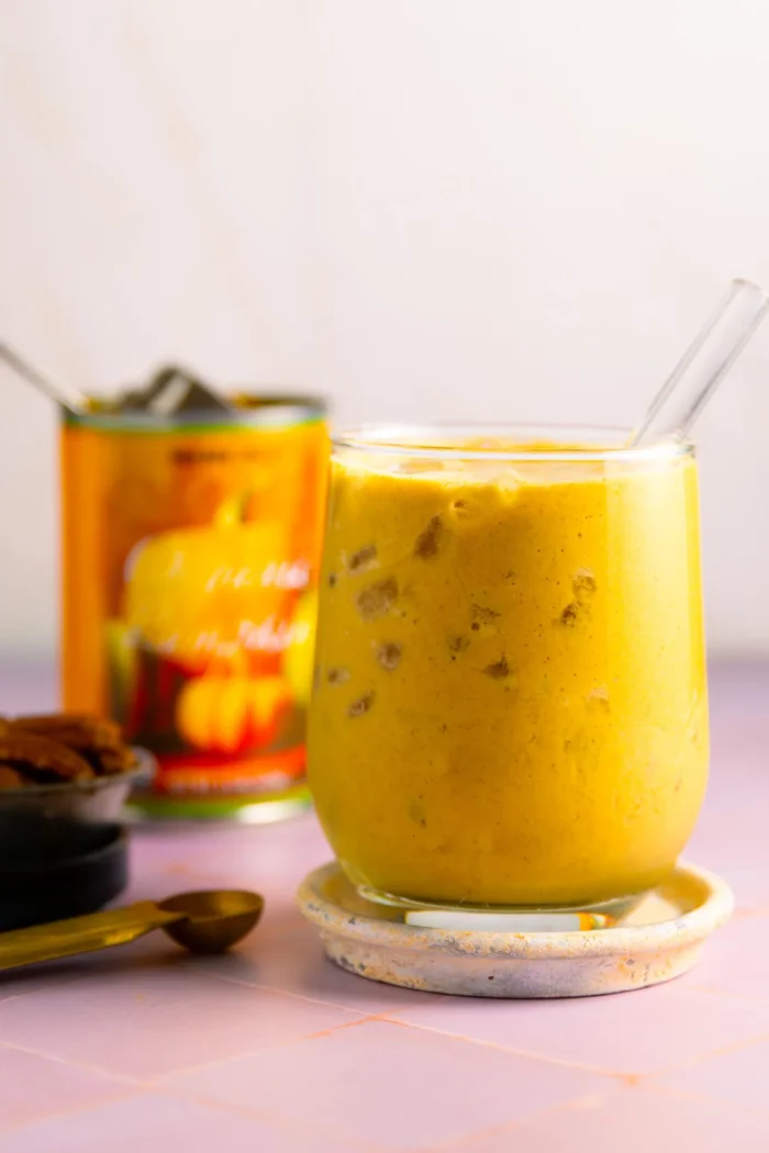 A glass of iced golden turmeric latte with a clear straw sits on a coaster. The yellow-orange drink resembles a Pumpkin Pie Protein Smoothie, with visible ice cubes. In the background, there’s a can with a pumpkin label—no banana in sight.