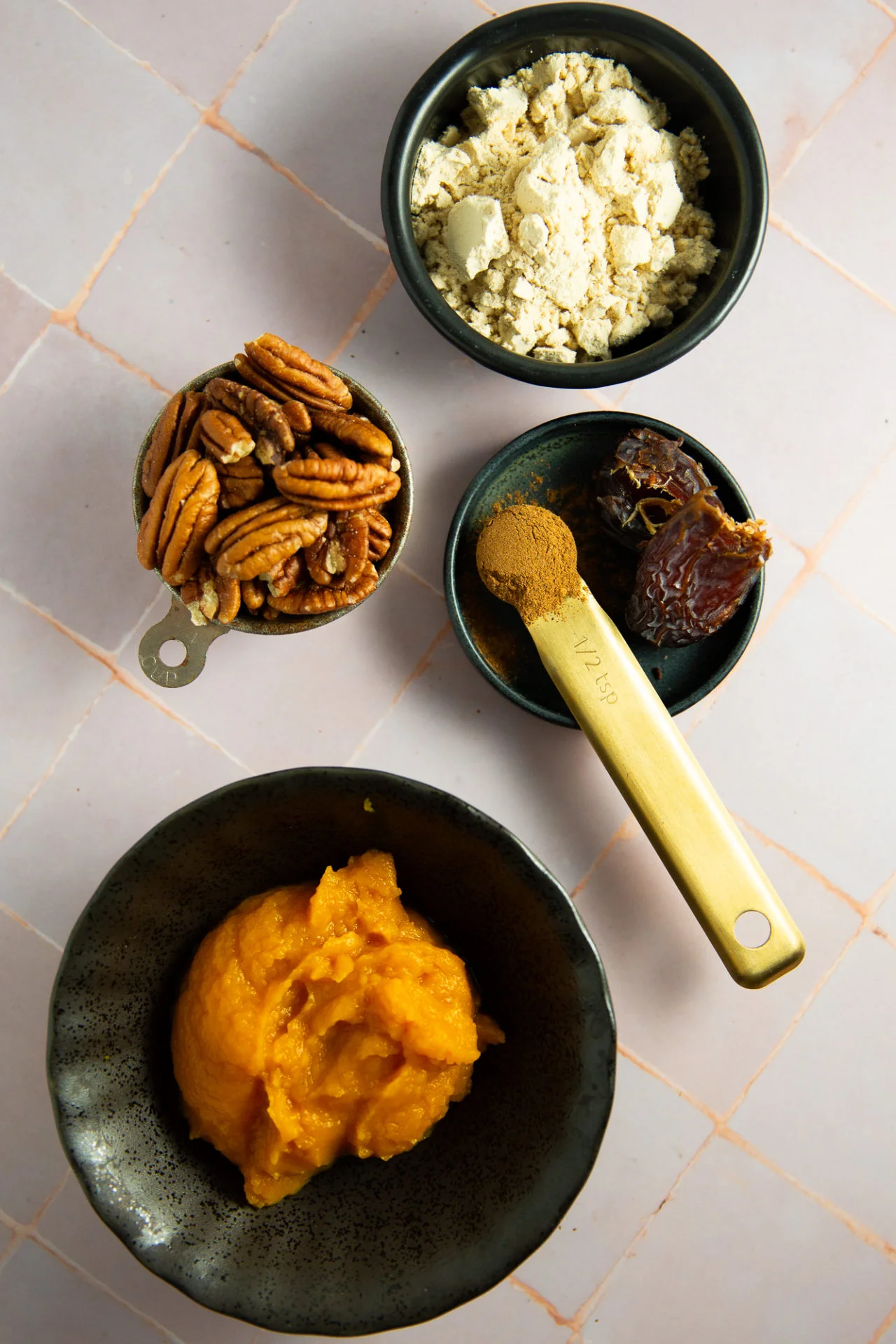 Four small bowls on a pink tiled surface hold ingredients for a pumpkin pie protein smoothie, no banana included: pecans, protein powder, pitted dates with cinnamon in a measuring spoon, and pumpkin puree. The bowls are assorted sizes and colors.