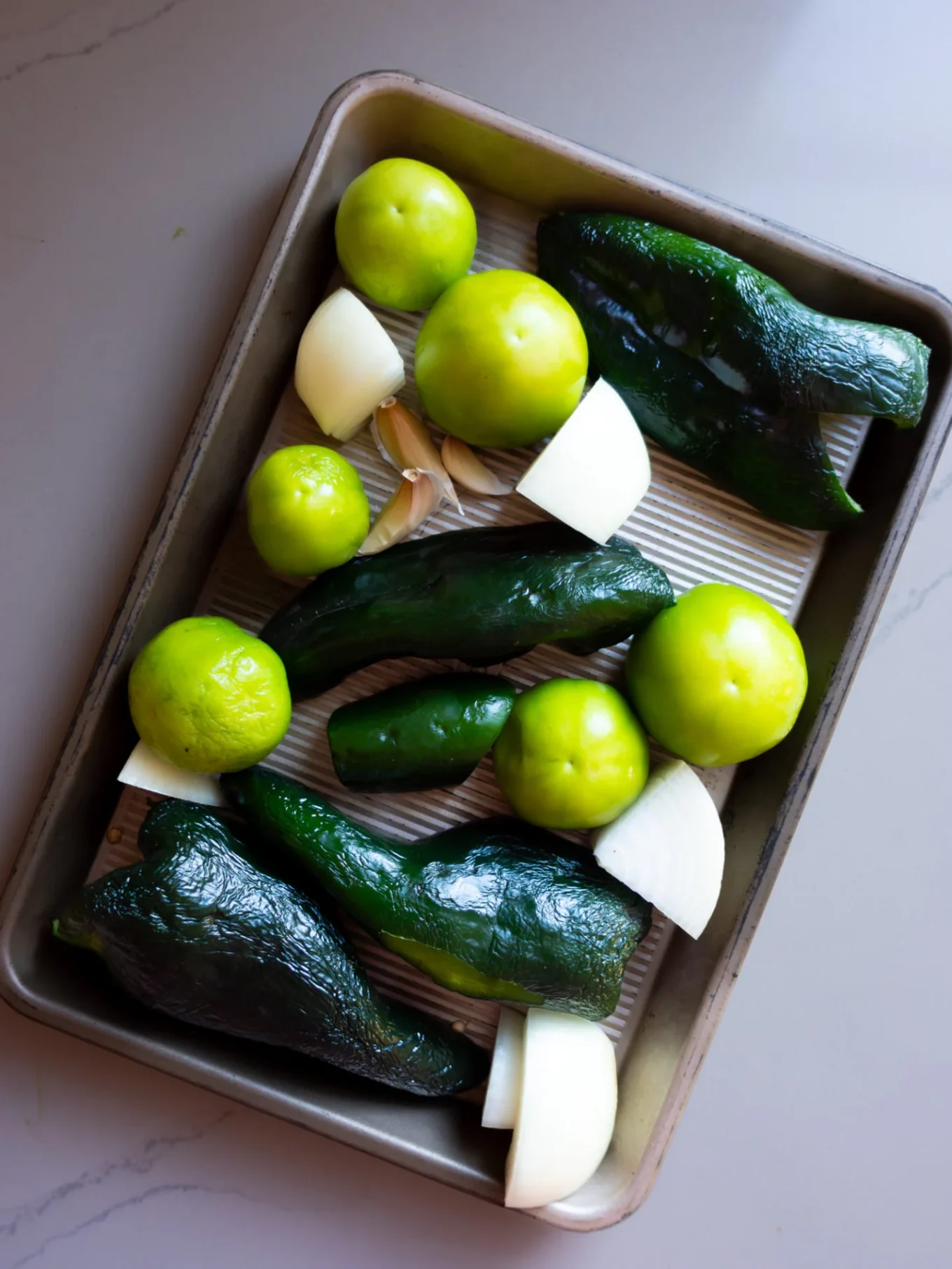 A baking sheet holds whole green poblano peppers, tomatillos with husks removed, quartered white onion pieces, and two garlic cloves—perfect for making a flavorful Roasted Poblano Salsa Verde—arranged on a light-striped metal tray atop a gray countertop.