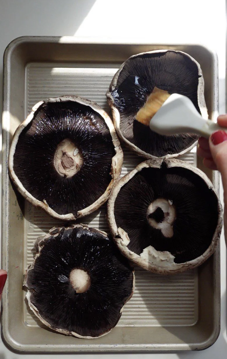 Four large portobello mushroom caps, ready for a Portobello Wellington, lie gills up on a metal baking tray. A hand brushes oil onto one, while sunlight highlights the mushrooms’ texture and the baking tray’s ridged surface.
