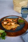A blue plate holds two crispy Shrimp Tacos with salsa, placed on a wooden tray. A small bowl of green sauce with a wooden spoon sits on the side. Lime wedges, cilantro, a glass of water, and a woven basket complete this authentic taco spread.