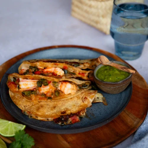 A blue plate holds two crispy Shrimp Tacos with salsa, placed on a wooden tray. A small bowl of green sauce with a wooden spoon sits on the side. Lime wedges, cilantro, a glass of water, and a woven basket complete this authentic taco spread.