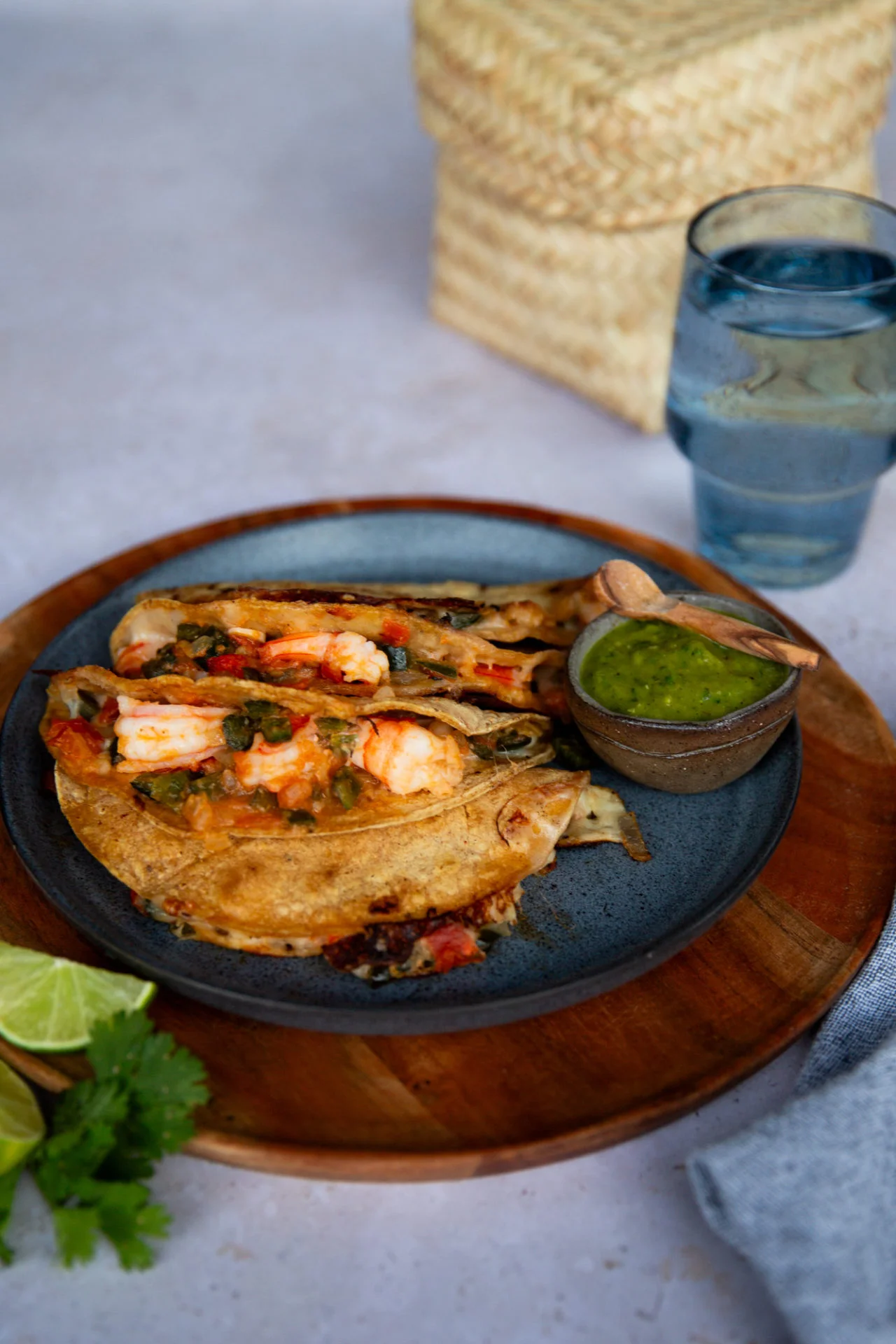 A blue plate holds two crispy Shrimp Tacos with salsa, placed on a wooden tray. A small bowl of green sauce with a wooden spoon sits on the side. Lime wedges, cilantro, a glass of water, and a woven basket complete this authentic taco spread.