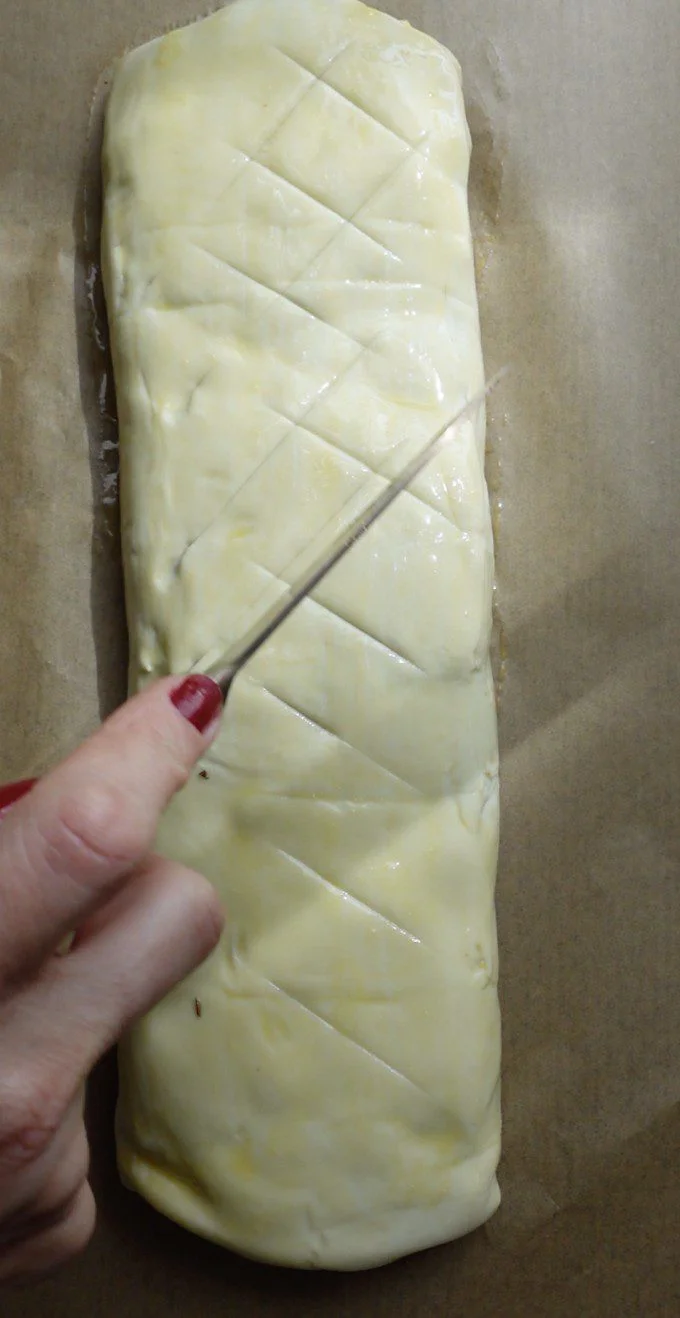 A hand with red nail polish scores diagonal lines onto a mushroom Wellington covered in egg wash, using a sharp knife; the pastry rests on a sheet of brown parchment paper.