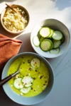 A bowl of creamy zucchini soup topped with cucumber slices and red pepper flakes sits on a white surface beside a bowl of sliced cucumbers and crumbled cheese, with a brown napkin and spoon nearby, all lit by natural sunlight.
