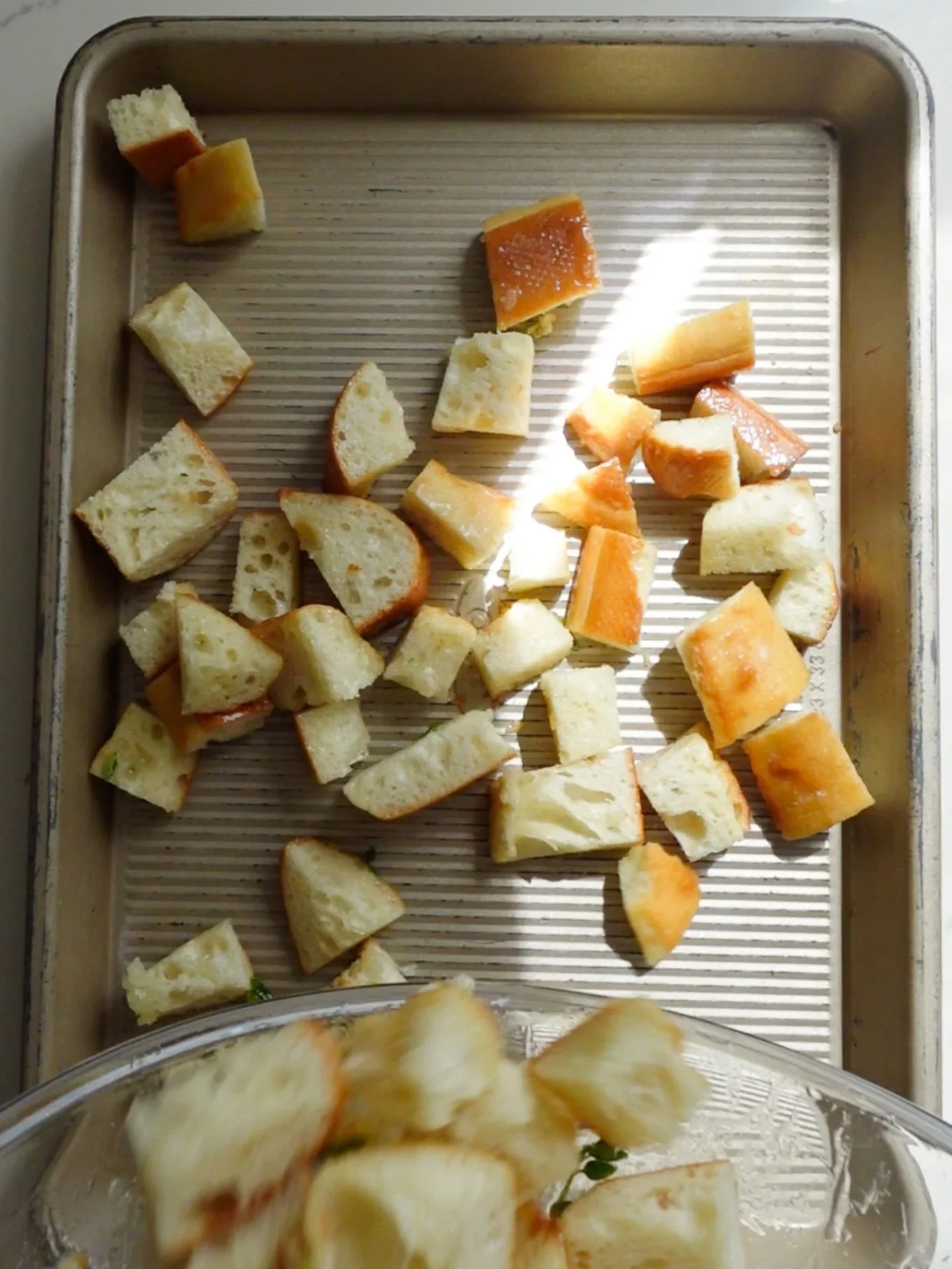 Cubed pieces of bread, perfect for Garlic Croutons, are scattered across a metal baking sheet. The ridged surface catches the light, highlighting their golden and white edges. A partially visible glass bowl with more bread cubes appears at the bottom.