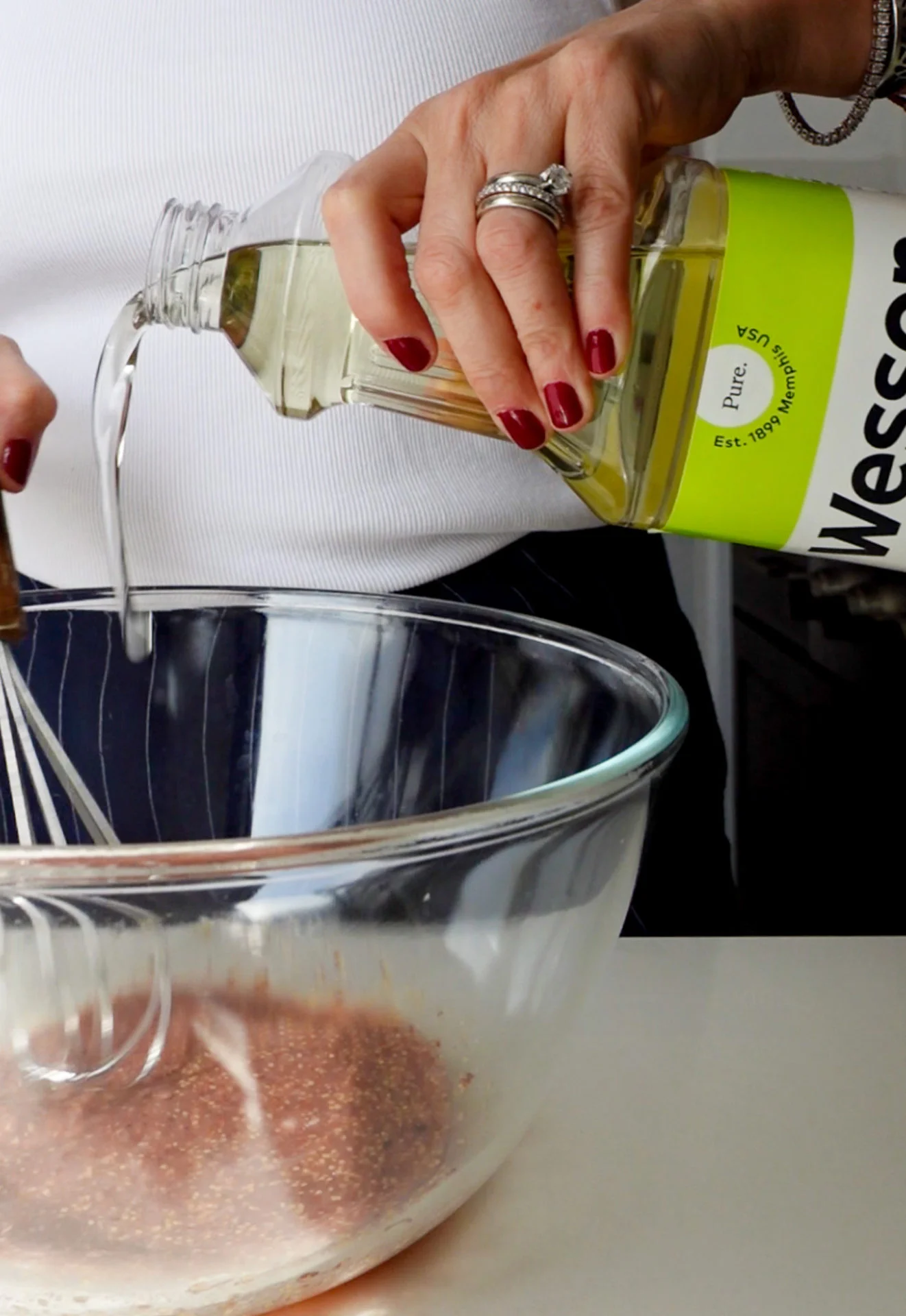 A person with red nail polish pours Wesson vegetable oil into a glass mixing bowl of brown powder, holding a whisk in the other hand—preparing to mix ingredients for a grainy mustard vinaigrette on a white countertop.