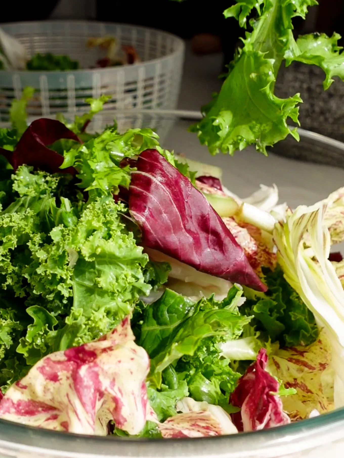 A close-up of fresh mixed salad greens, including curly kale, red radicchio, and frisée, in a glass bowl. The vibrant greens and purples are crisp and leafy. In the background, Garlic Croutons and a salad spinner rest on the kitchen counter.