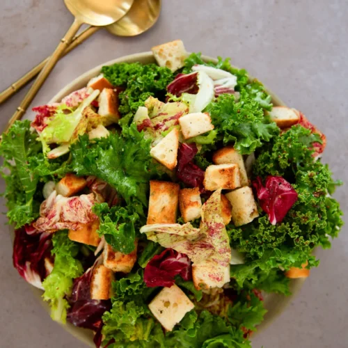 A bowl of fresh green and purple Winter Green Salad with crunchy garlic croutons on top sits on a gray surface. Two gold salad servers are placed beside the bowl. The salad appears vibrant and crisp.