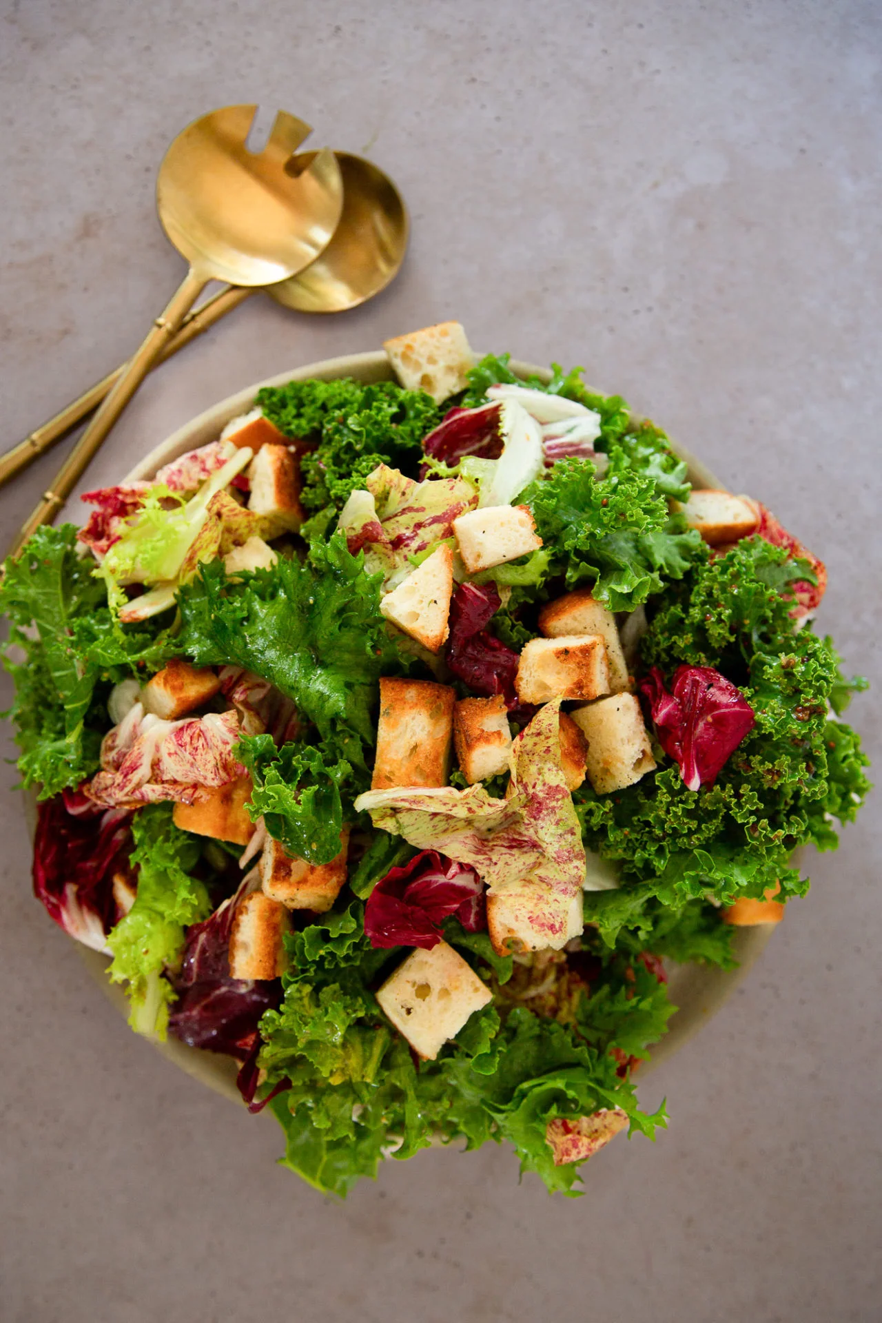 A bowl of fresh green and purple Winter Green Salad with crunchy garlic croutons on top sits on a gray surface. Two gold salad servers are placed beside the bowl. The salad appears vibrant and crisp.