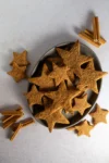 A gray oval plate holds star-shaped Hojarascas, traditional Mexican cinnamon cookies dusted with cinnamon. Several cookies are scattered around the plate, while bundles of whole cinnamon sticks add a rustic, festive touch to the scene.