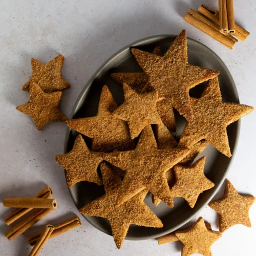 A gray oval plate holds star-shaped Hojarascas, traditional Mexican cinnamon cookies dusted with cinnamon. Several cookies are scattered around the plate, while bundles of whole cinnamon sticks add a rustic, festive touch to the scene.
