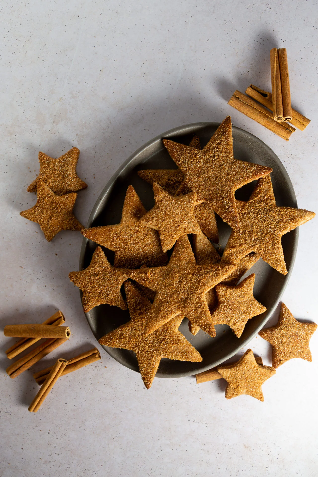 A gray oval plate holds star-shaped Hojarascas, traditional Mexican cinnamon cookies dusted with cinnamon. Several cookies are scattered around the plate, while bundles of whole cinnamon sticks add a rustic, festive touch to the scene.