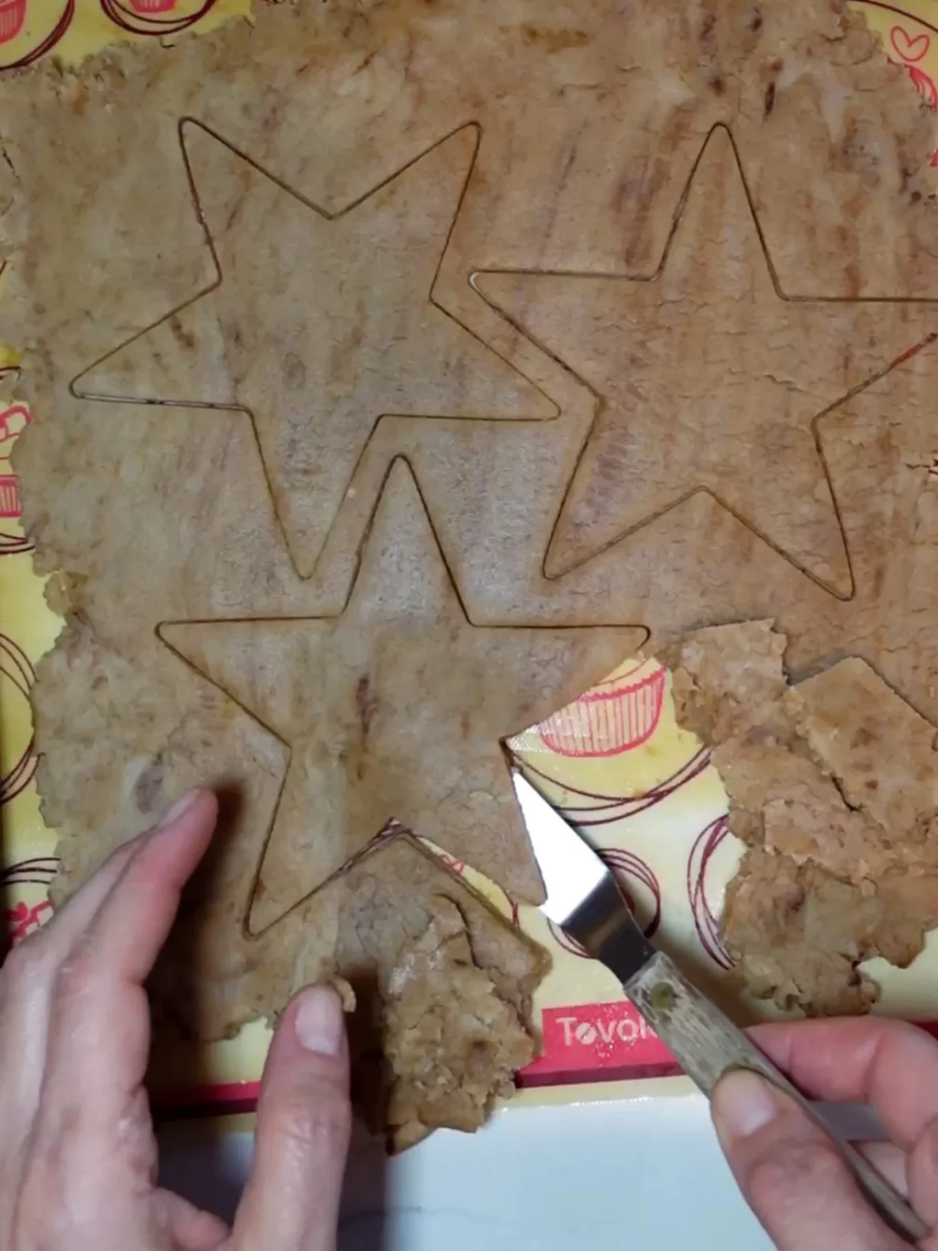 A pair of hands uses a knife to cut star shapes out of brown dough for Hojarascas, traditional Mexican cookies coated in cinnamon sugar, on a yellow baking mat decorated with red drawings of rolling pins, whisks, and cupcakes.
