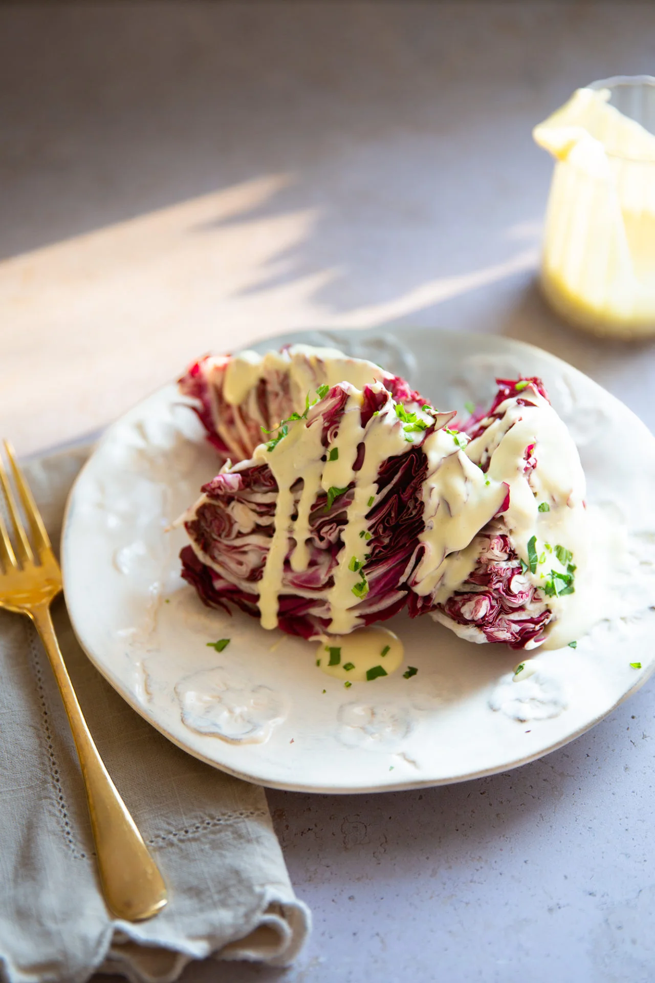 A plate with wedges of purple radicchio drizzled with bold, Creamy Parmesan Dressing and garnished with chopped herbs. A gold fork and beige napkin sit beside the plate, while more dressing glows in soft natural light. Easy elegance on display.