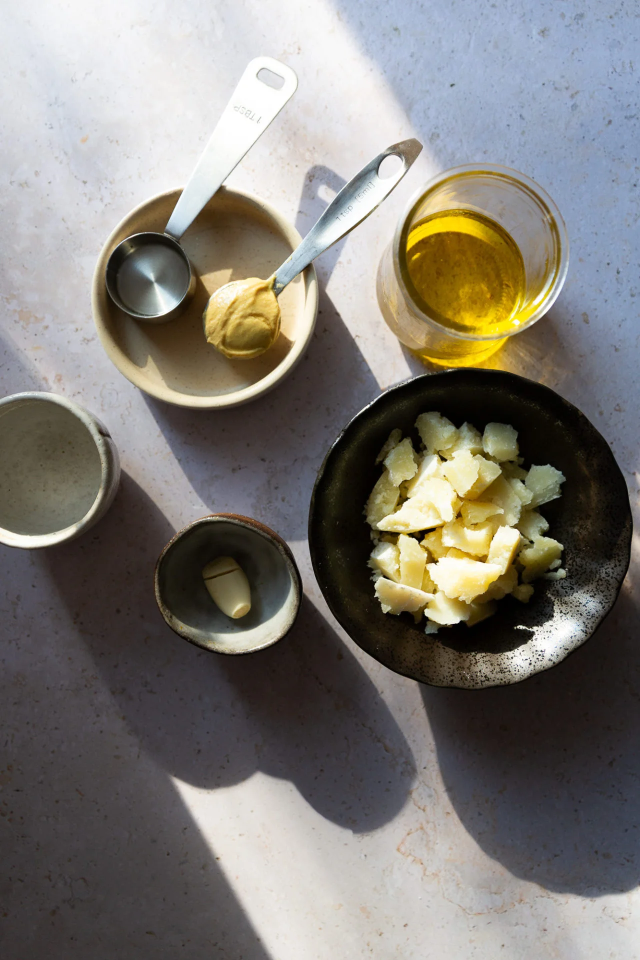 A flat lay of ingredients for an easy creamy parmesan dressing: a black bowl with shaved parmesan, bold Dijon mustard on a beige plate, olive oil in a glass, garlic in a white bowl, and measuring spoons—all bathed in sunlight.