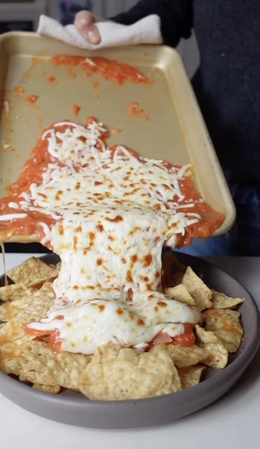 A hand tips a baking sheet, sliding a layer of melted cheese and tomato sauce onto tortilla chips, creating gooey Chilaquiles Rojos. The cheese is golden-brown and stretchy, making this dish a delicious chilaquiles recipe you&rsquo;ll crave.