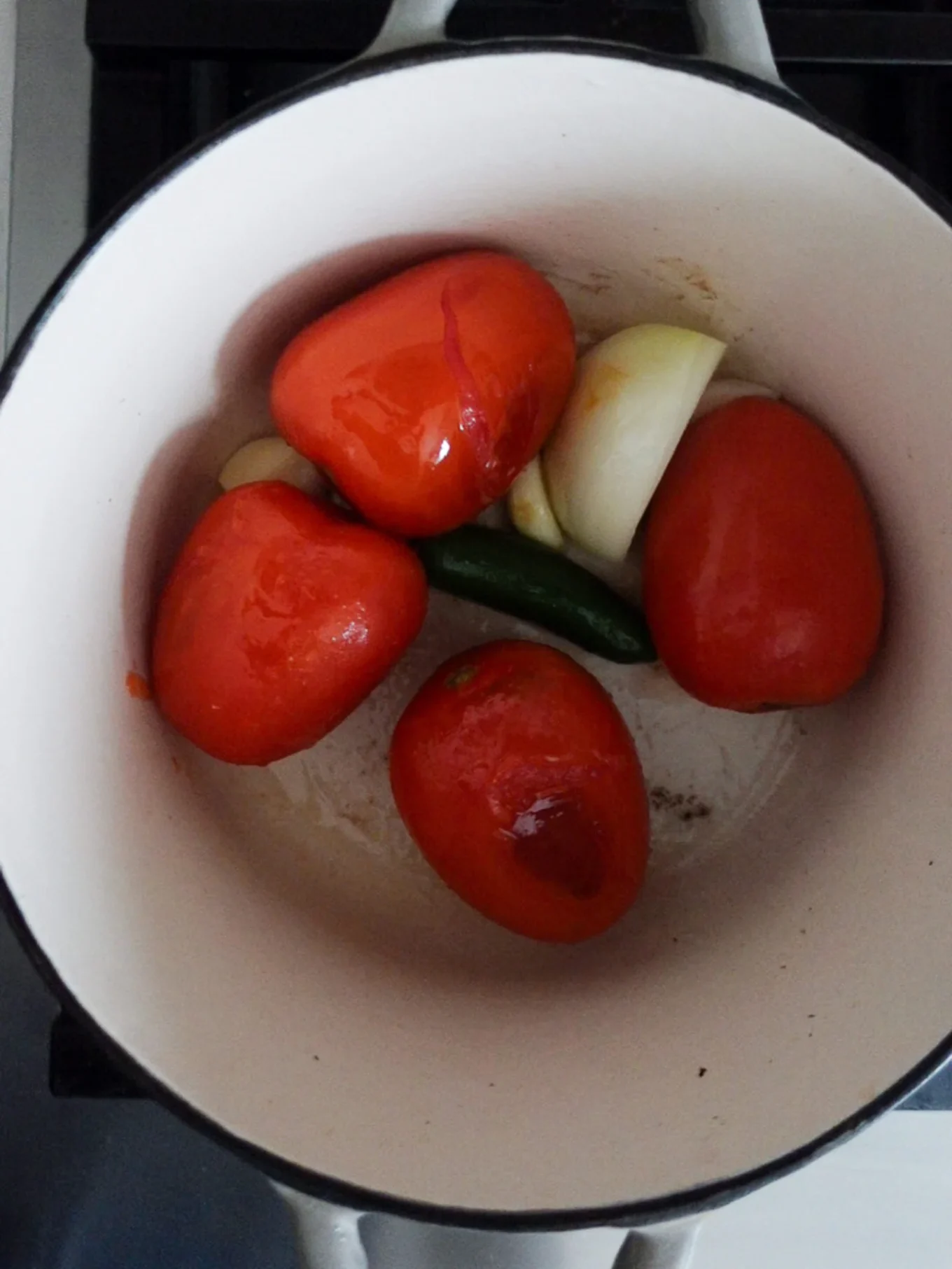 Cinco tomates rojos Roma, un chile verde entero, varios dientes de ajo y un trozo de cebolla blanca se acomodan en una olla blanca sobre la estufa-frescos y sin cortar, listos para que este m&eacute;todo &uacute;nico de chilaquiles cree la base perfecta para los Chilaquiles Rojos.