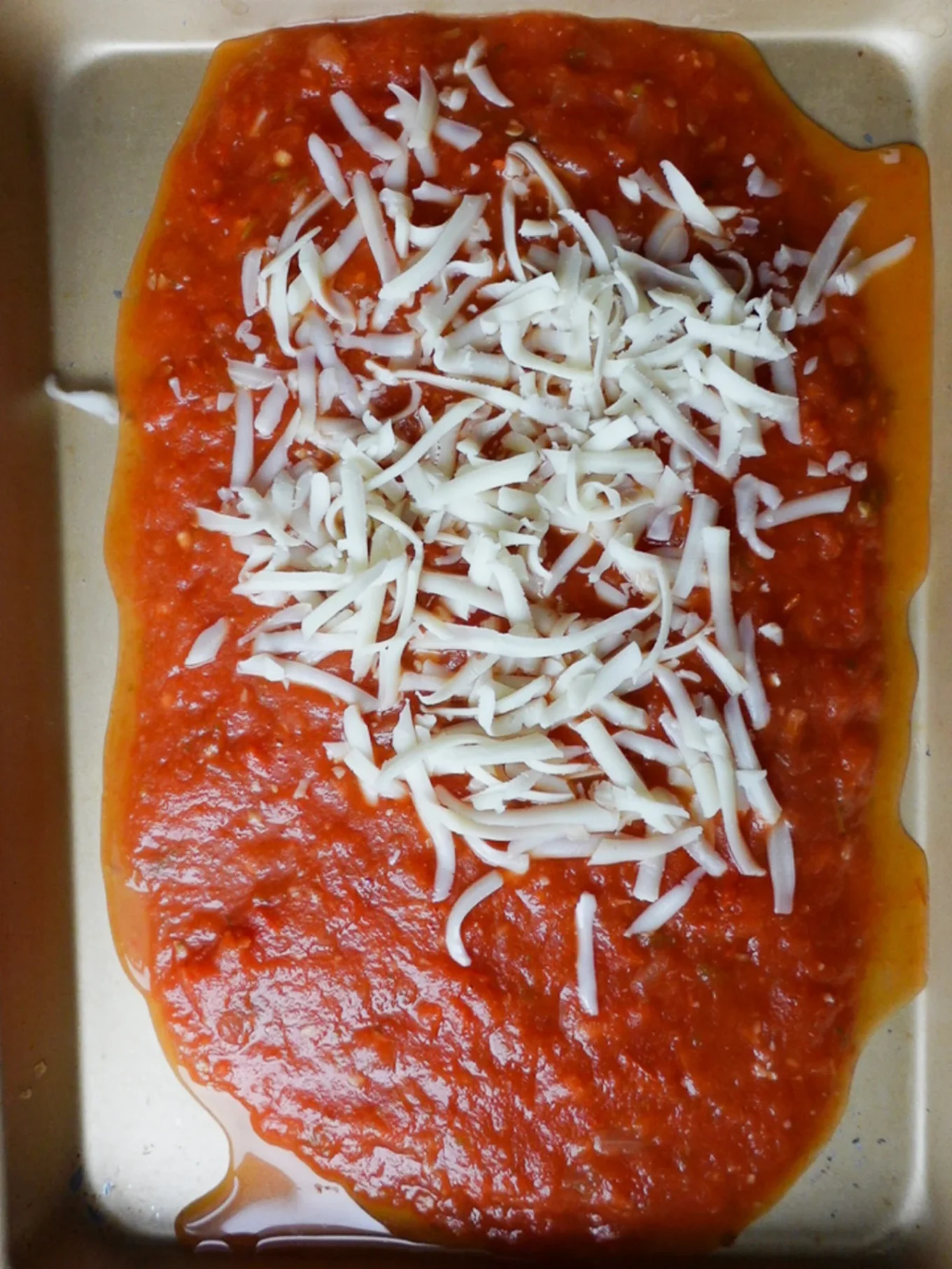 A close-up of a baking tray containing a layer of tomato sauce topped with a generous amount of shredded white cheese, perfect for a delicious chilaquiles recipe. The sauce spreads unevenly, with some oil pooling around the edges.