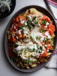 A plate of Chilaquiles Rojos topped with red salsa, melted cheese, sliced onions, chopped cilantro, and cream. Crispy tortilla chips peek through. A bowl of onions and a striped cloth napkin are nearby on a white surface.