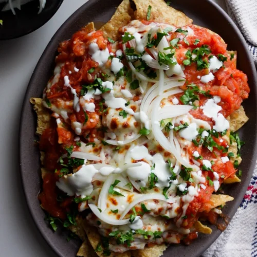 A plate of Chilaquiles Rojos topped with red salsa, melted cheese, sliced onions, chopped cilantro, and cream. Crispy tortilla chips peek through. A bowl of onions and a striped cloth napkin are nearby on a white surface.