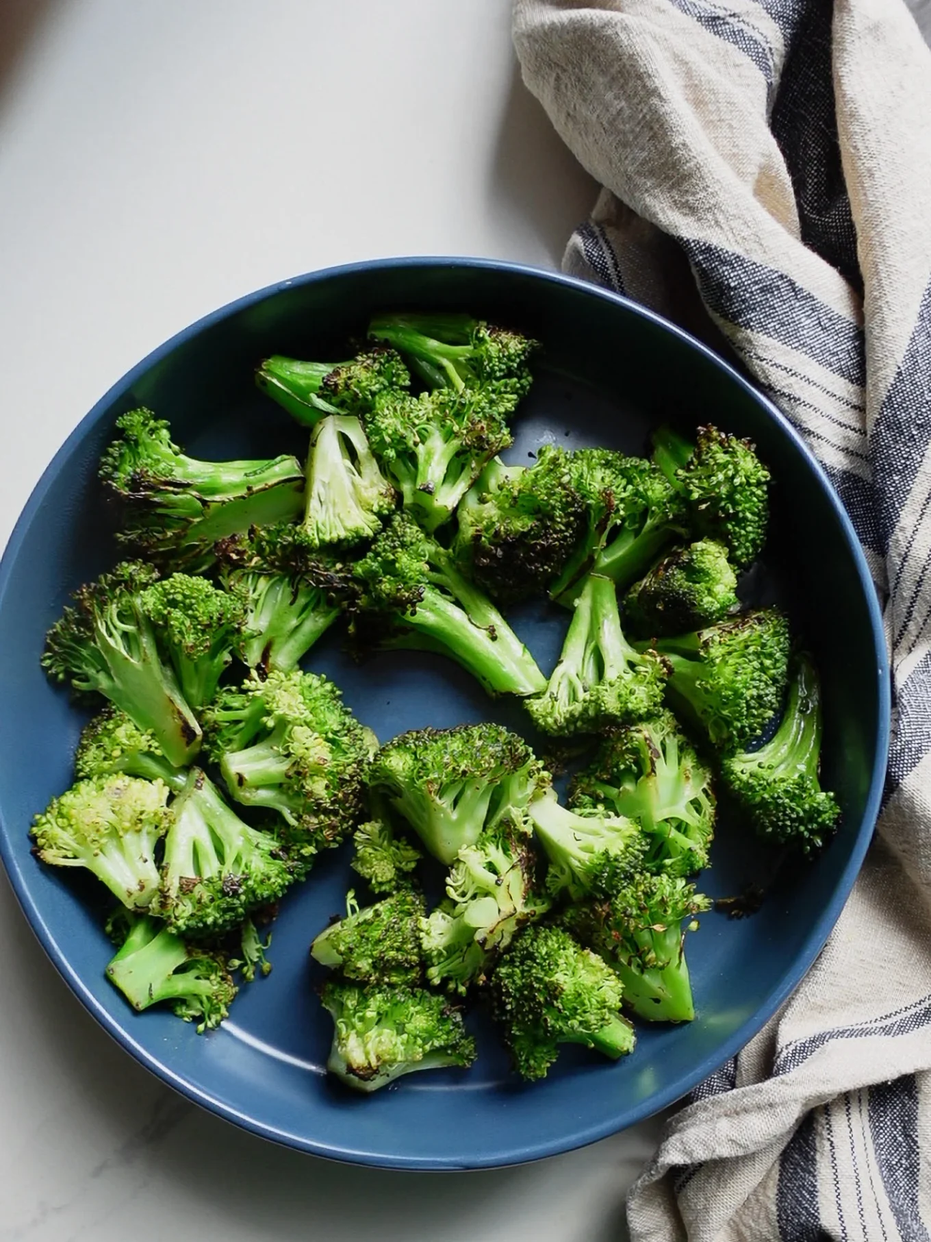 A blue bowl filled with roasted broccoli florets sits on a light surface, showcasing this delicious Roasted Broccoli Salad. The broccoli is slightly browned and seasoned. A striped cloth napkin in beige and navy rests nearby on the right side of the image.