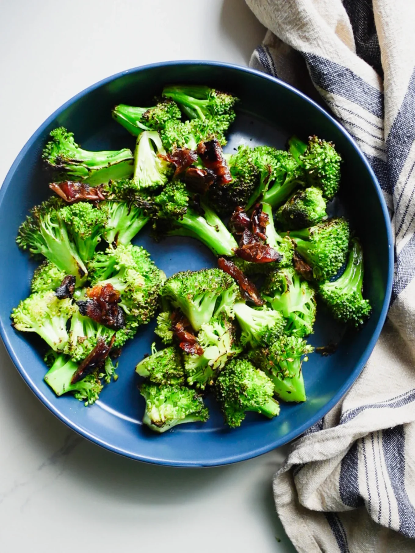 A blue plate of Roasted Broccoli Salad with caramelized onions sits on a light surface. A beige and navy striped cloth napkin is partially visible beside the plate. The salad features lightly browned, fresh broccoli florets.