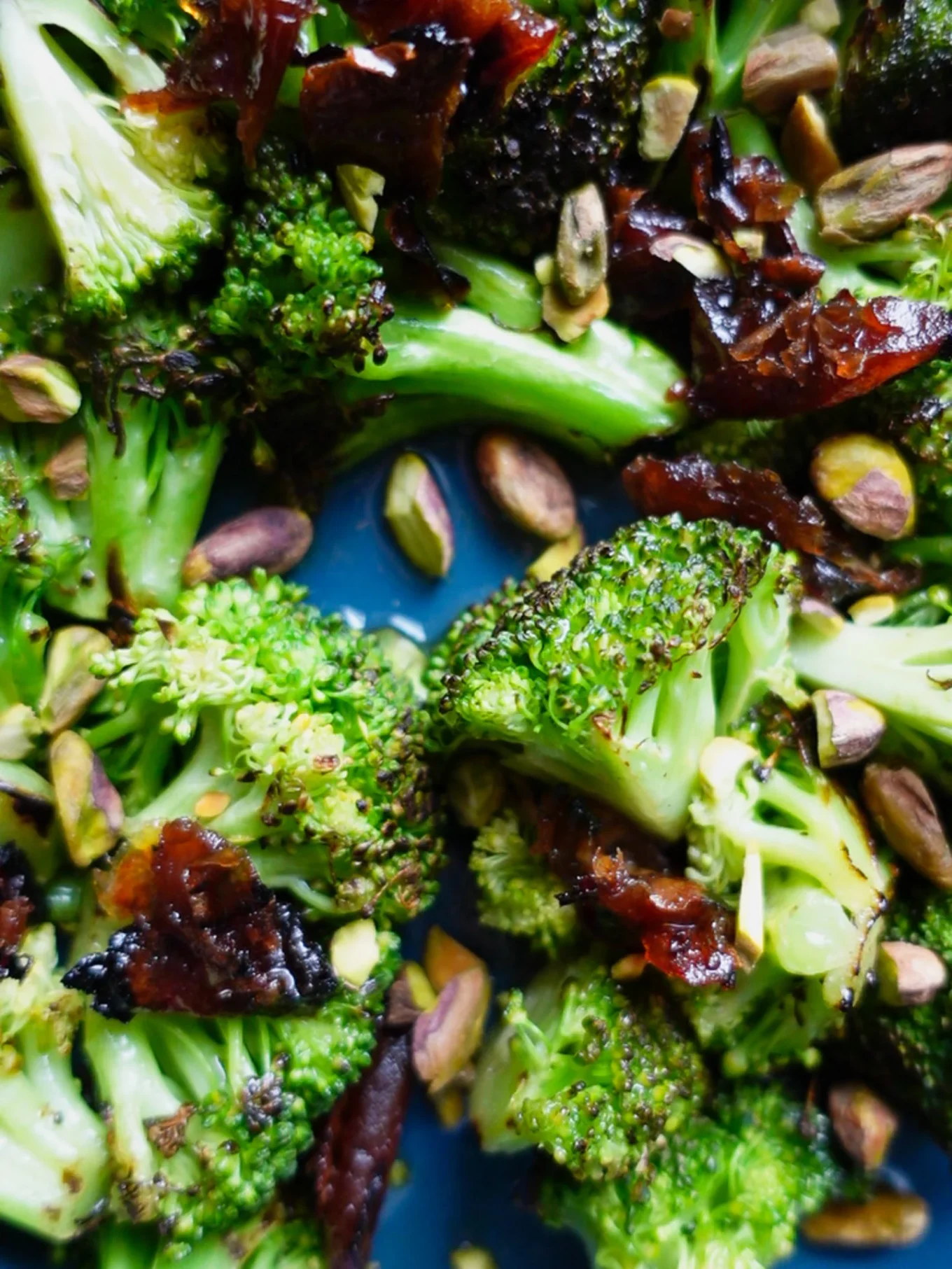 Close-up of Charred broccoli salad mixed with crispy bacon, whole pistachios, and sweet dates, all on a blue surface. The roasted-style broccoli is charred in places, adding texture and color contrast to the vibrant green.