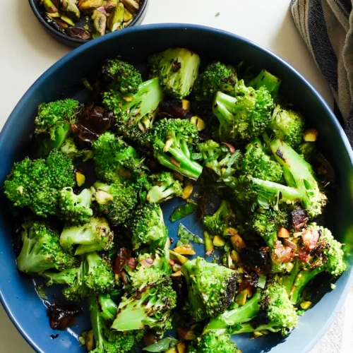 A blue bowl filled with roasted broccoli florets, chopped pistachios, and pieces of what appears to be caramelized onions or dried fruit makes this Charred Broccoli Salad shine. A small dish of more pistachios and a striped towel sit nearby on a white surface.