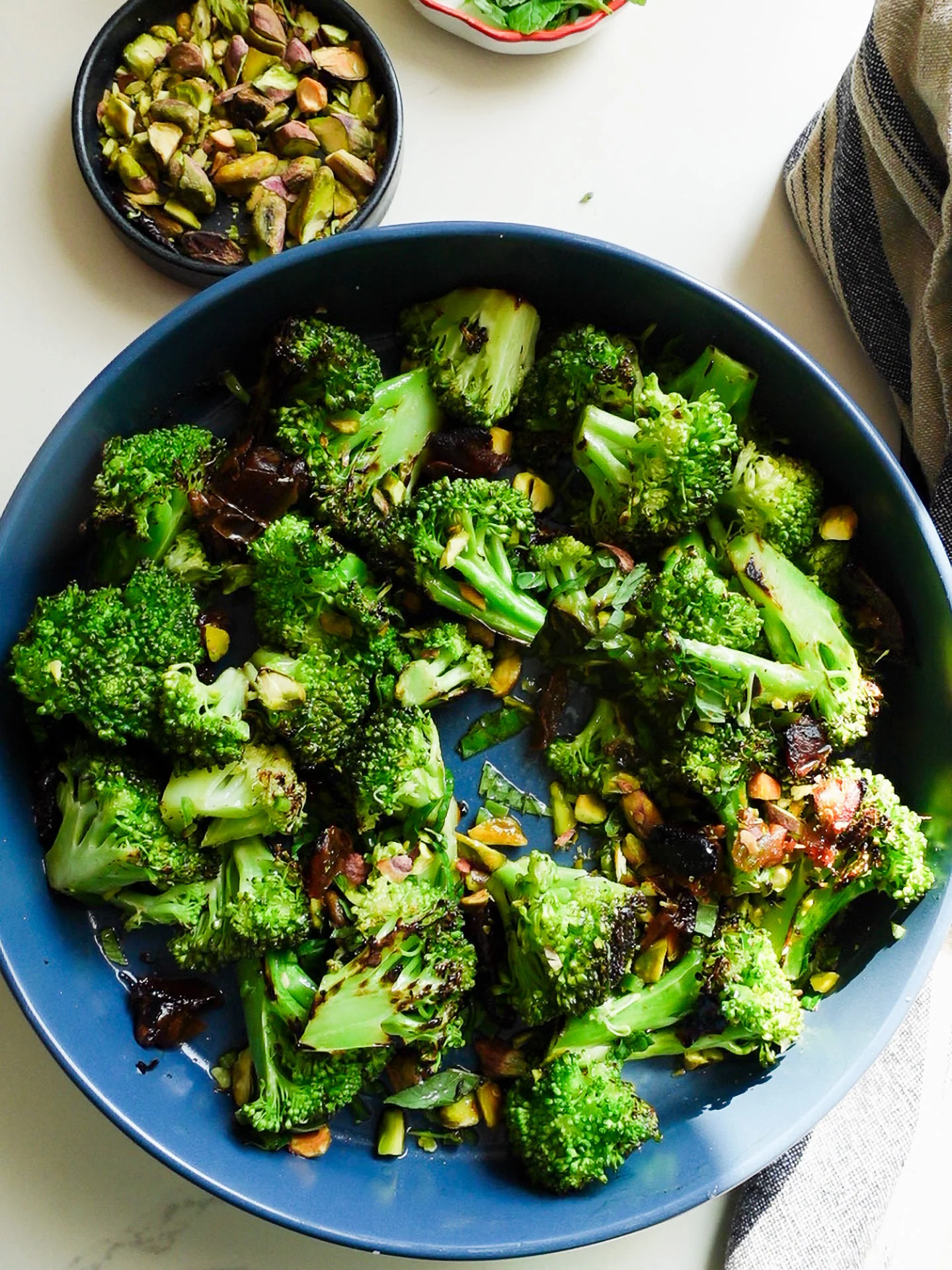 A blue bowl filled with vibrant Roasted Broccoli Salad, tossed with chopped pistachios and sweet roasted dates. A small bowl of pistachios and a striped napkin sit nearby on the white surface, enhancing the fresh presentation.