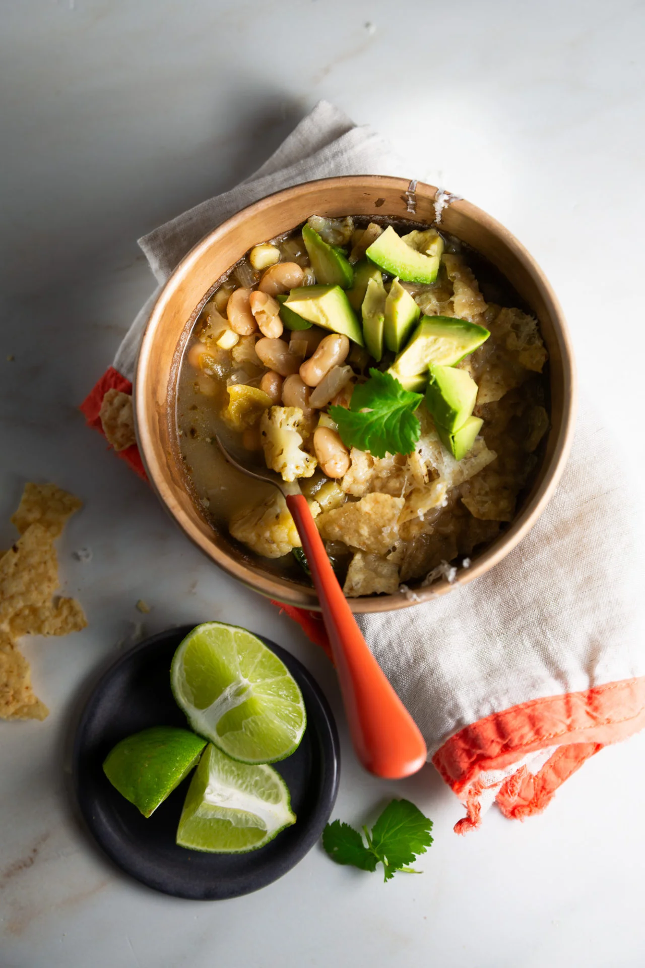 A bowl of Creamy White Bean Chili with chunks of avocado and cilantro garnish sits on a napkin with an orange border. Nearby, lime wedges and cilantro rest on a dark plate, while broken tortilla chips are scattered on white marble.