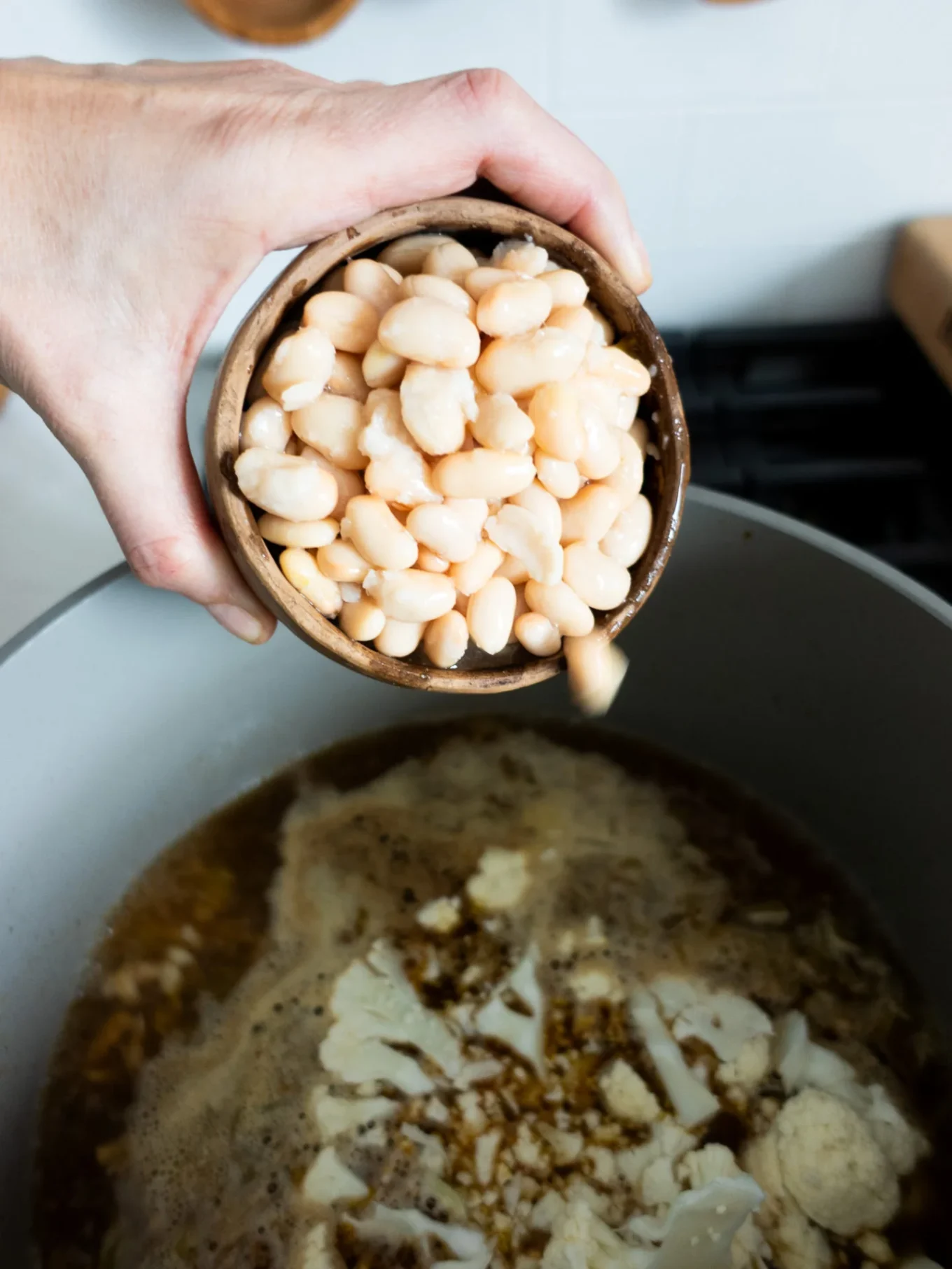 A hand holds a small wooden bowl of white beans above a large pot of simmering broth with oil, seasonings, and onion, preparing to pour the beans into the pot for a flavorful White Bean Chili.