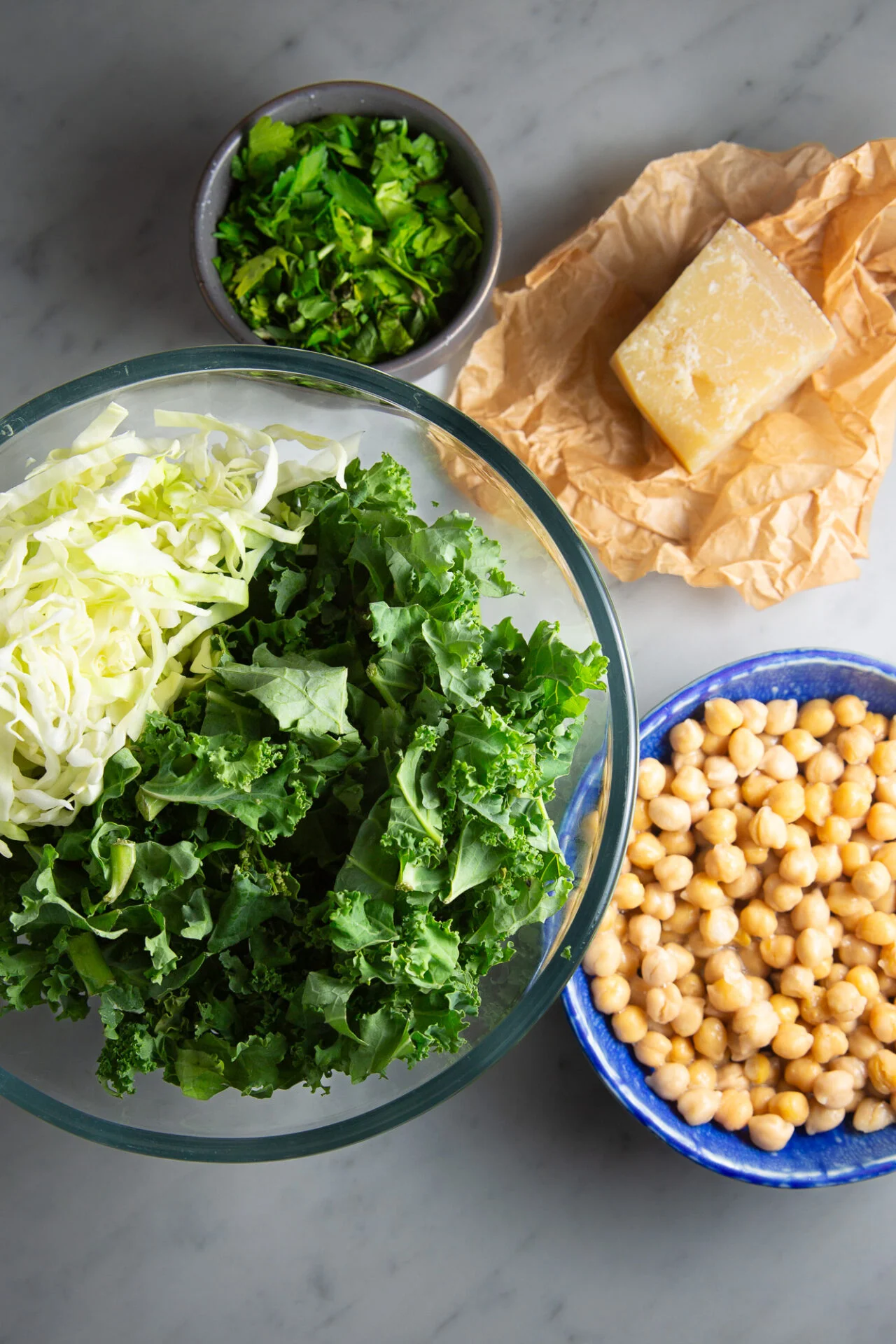 A top-down view of a marble surface displays a glass bowl of chopped kale and cabbage, a blue bowl of chickpeas, a small bowl of herbs, and Parmesan cheese&mdash;perfect ingredients for recreating the Hillstone Emerald Kale Salad at home.