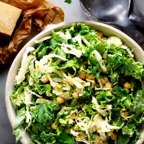 A bowl of Emerald Kale Salad with shredded cabbage, chickpeas, and herbs sits on a marble surface. A block of Parmesan cheese on crumpled brown paper and black salad servers are nearby, creating a bright and fresh Hillstone-inspired scene.