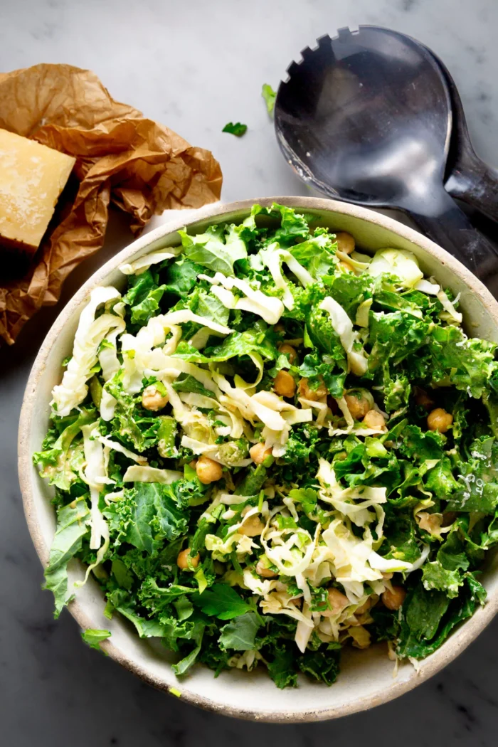 A bowl of Emerald Kale Salad with shredded cabbage, chickpeas, and herbs sits on a marble surface. A block of Parmesan cheese on crumpled brown paper and black salad servers are nearby, creating a bright and fresh Hillstone-inspired scene.