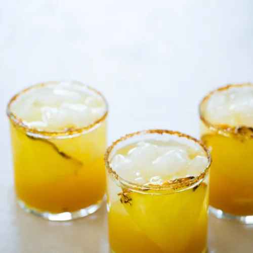 Three short glasses filled with a yellow-orange drink and ice cubes are arranged on a light surface. Each glass has a chili powder and salt rim, and visible chili peppers float inside the beverages.