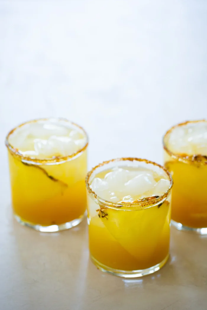 Three short glasses filled with a yellow-orange drink and ice cubes are arranged on a light surface. Each glass has a chili powder and salt rim, and visible chili peppers float inside the beverages.