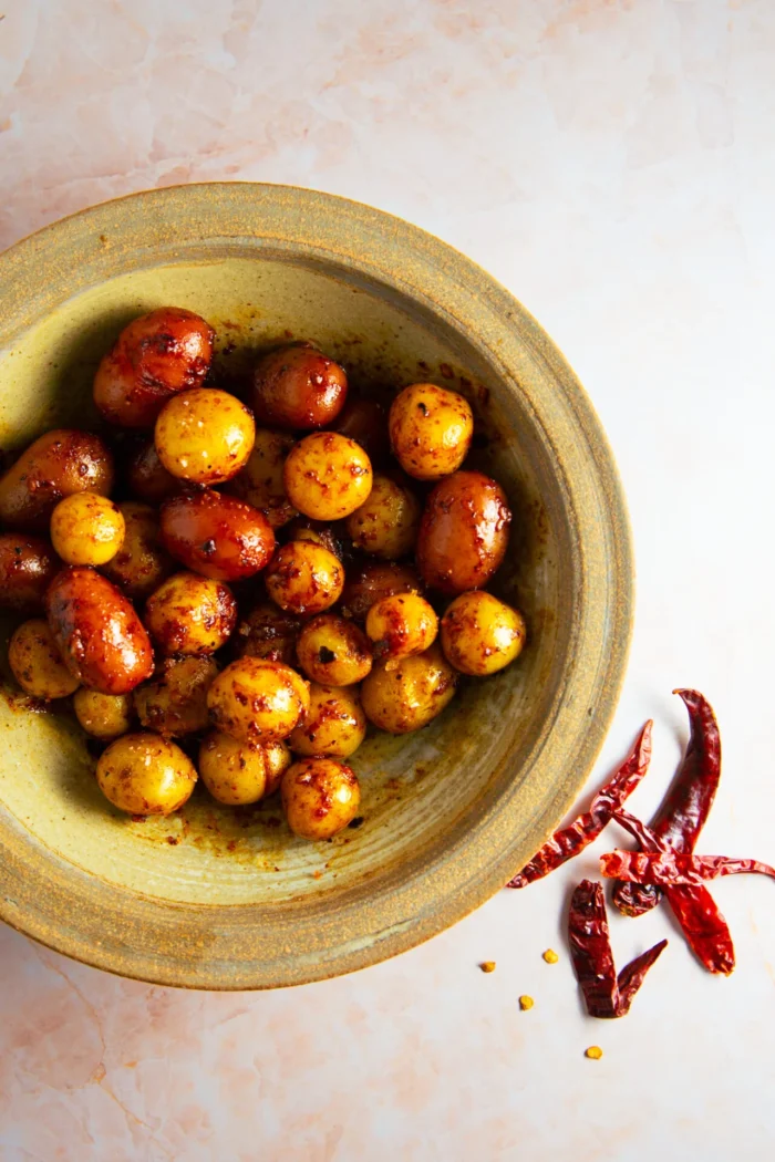 A rustic green bowl holds Spicy Mexican Potatoes&mdash;baby potatoes coated in a reddish seasoning. Next to the bowl on a pale surface are whole dried red chilies and scattered chili seeds, hinting at their fiery flavor.
