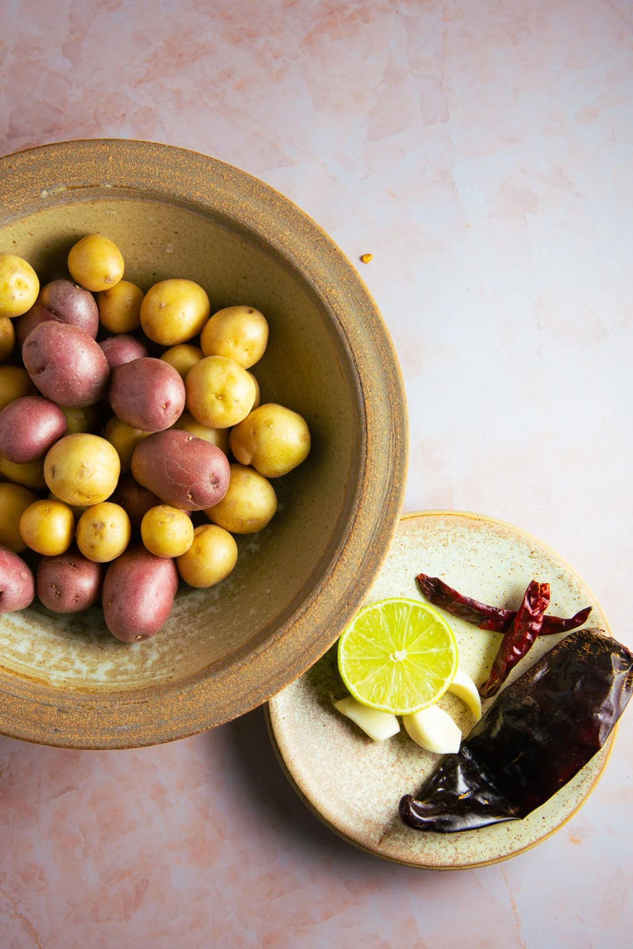 A rustic bowl of small red and yellow potatoes—perfect for Spicy Mexican Potatoes—sits on a pink marble surface. Nearby, a plate displays a halved lime, garlic cloves, dried red chilies, and a dark chili pepper for Papas a la Diabla.