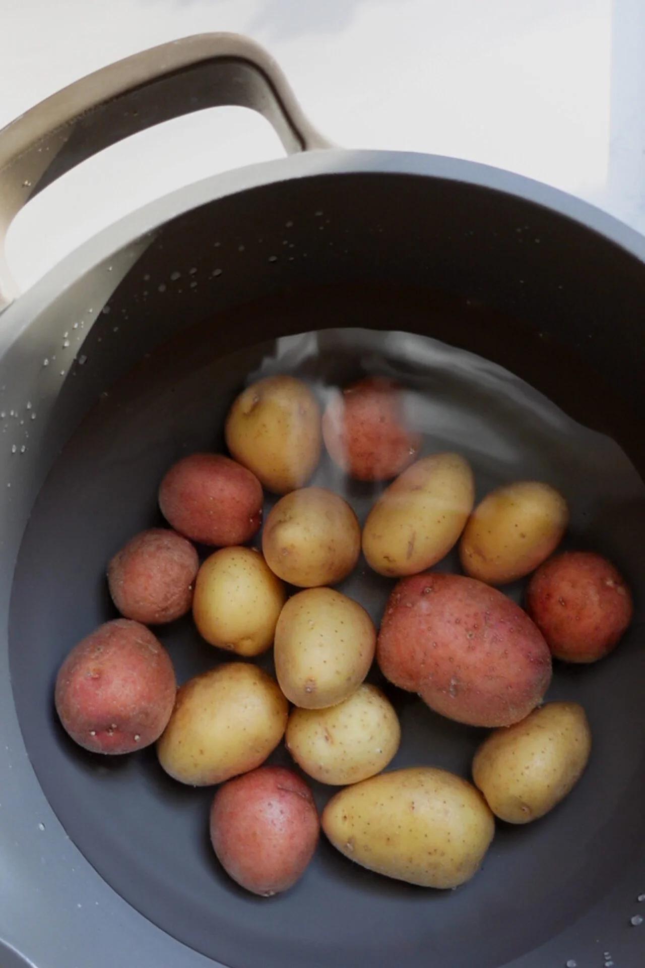 A large pot filled with water contains several unpeeled small red and yellow potatoes, perfect for a creamy potato salad. The potatoes are mostly submerged, light reflects off the water surface, and a wide-handled gray pot sits at the top left.