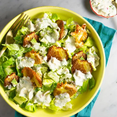 A green bowl of crispy potato salad features smashed potatoes atop chopped romaine, all drizzled with creamy ranch dressing. A gold fork rests in the bowl, with extra ranch in a small dish nearby on a teal napkin over marble.