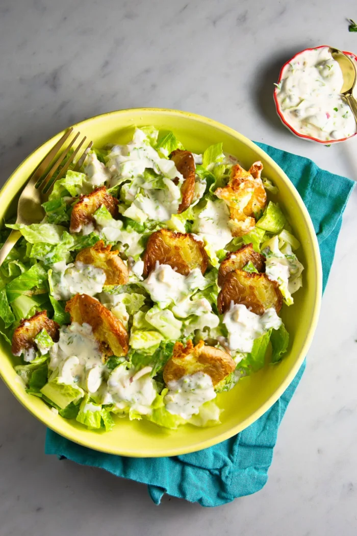 A green bowl of crispy potato salad features smashed potatoes atop chopped romaine, all drizzled with creamy ranch dressing. A gold fork rests in the bowl, with extra ranch in a small dish nearby on a teal napkin over marble.