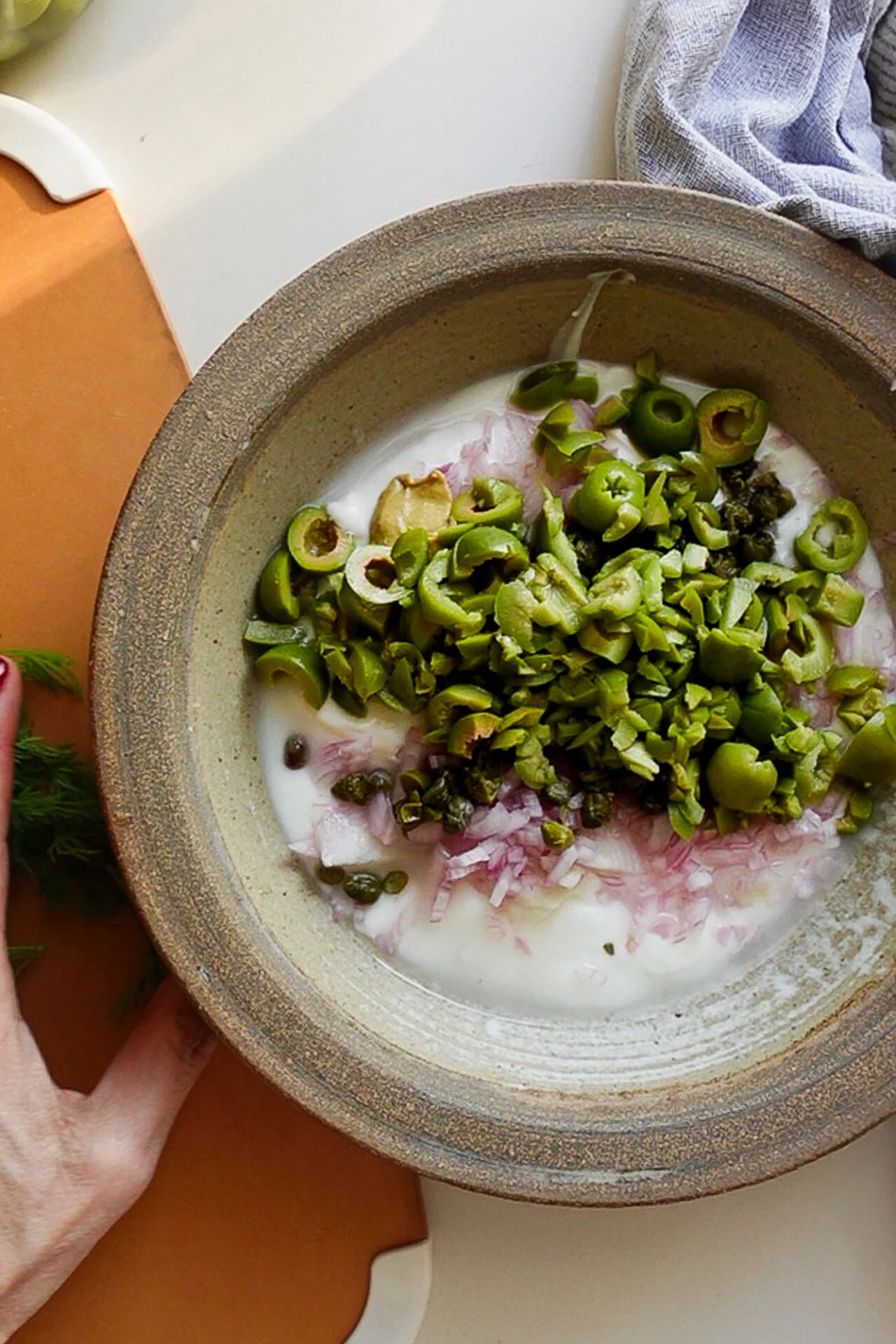 A ceramic bowl on a countertop holds toppings for a creamy potato salad: yogurt, finely chopped red onions, sliced green olives, and capers. A hand touches the bowl's edge; fresh dill peeks from the left corner beside a light purple cloth.