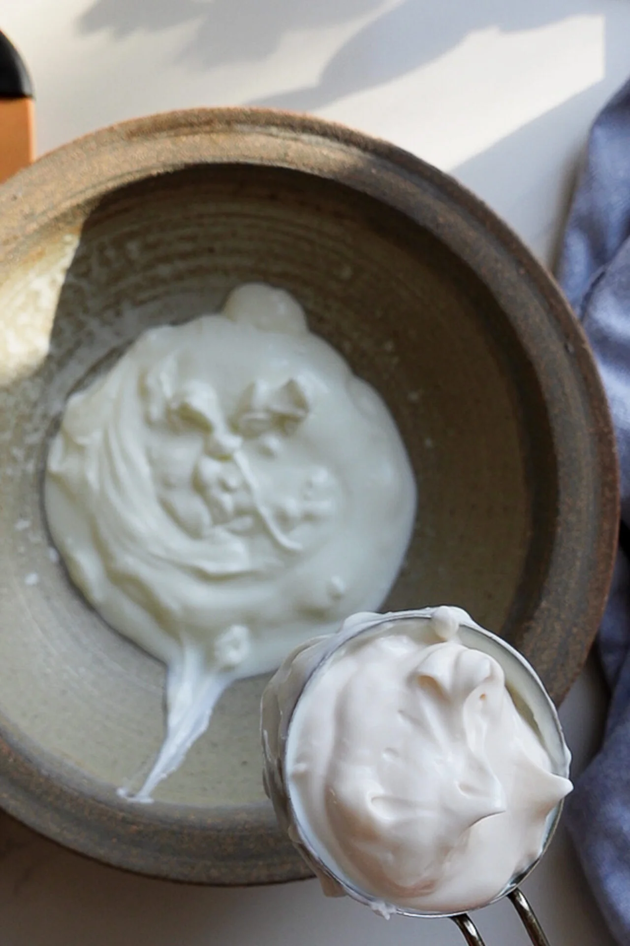 A brown ceramic bowl contains a dollop of thick, white whipped cream or yogurt&mdash;perfect for making creamy potato salad. A metal measuring cup in the foreground holds more of the creamy base, all softly lit by natural light from above.