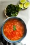 A blue bowl of steaming Caldo de Repollo, a Mexican cabbage soup with a cilantro garnish, sits on a white surface. Nearby are a small bowl of fresh cilantro leaves, four lime wedges on a plate, and a striped cloth in the corner.